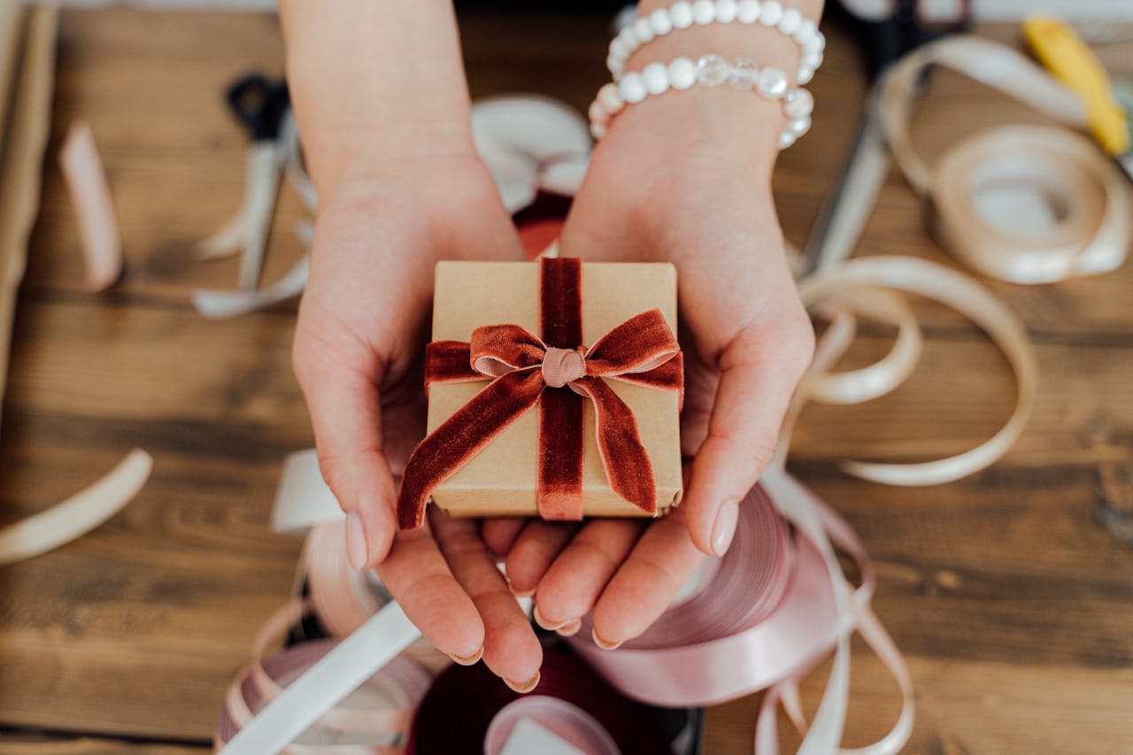 Hands presenting a small gift box wrapped with red velvet ribbon, surrounded by crafting materials.